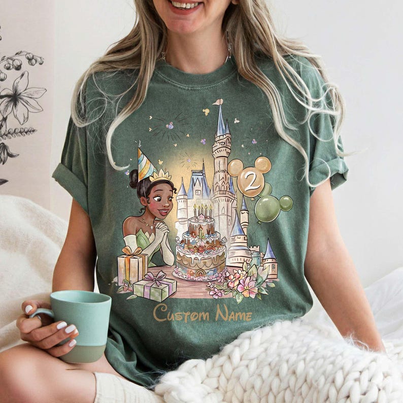May include: Green t-shirt with a cartoon illustration of a princess celebrating a birthday with a castle, cake, and balloons. The design includes the text "Custom Name" below the illustration. The shirt is worn by a person holding a blue mug.