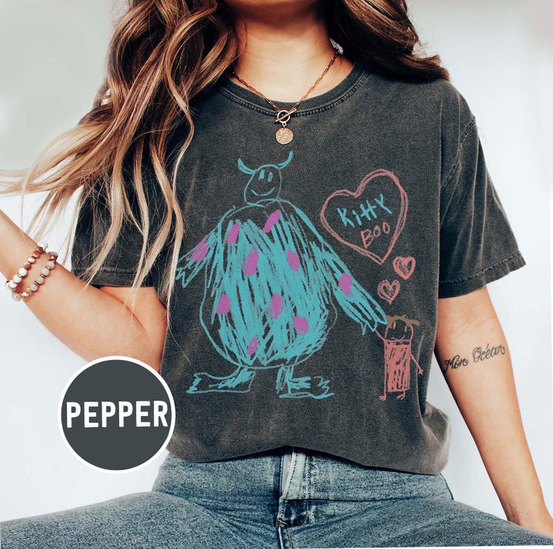 May include: Dark gray t-shirt featuring a whimsical drawing of a blue monster with purple spots, a pink heart with "Kitty Boo" text, and small pink hearts. The design is in a crayon-like style.