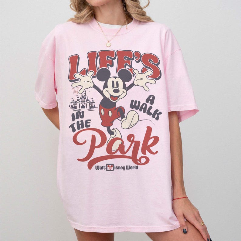 May include: A light pink t-shirt featuring a graphic of Mickey Mouse with the text "Life's a Walk in the Park" in red and white. The Walt Disney World logo is at the bottom. The shirt has short sleeves.