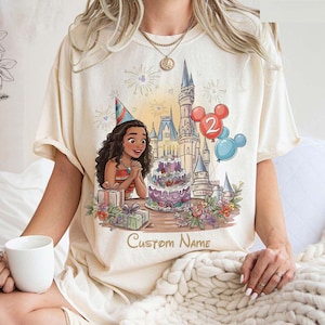 May include: Cream t-shirt with a birthday design. It features a cartoon character, a castle, a cake, and balloons. The design includes the text "Custom Name". The character wears a party hat and is surrounded by gifts and flowers.