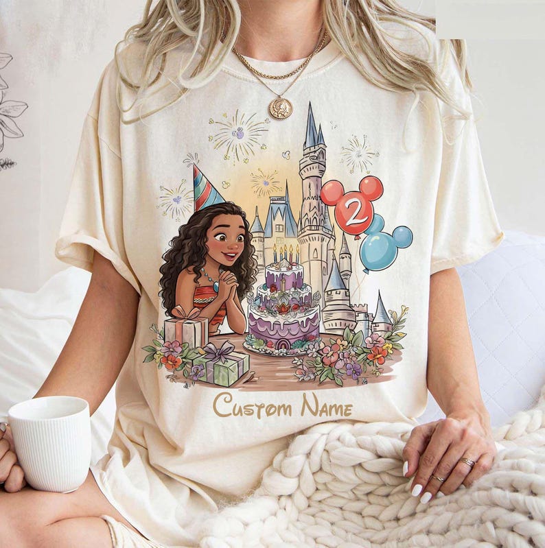 May include: Cream t-shirt with a birthday design. It features a cartoon character, a castle, a cake, and balloons. The design includes the text "Custom Name". The character wears a party hat and is surrounded by gifts and flowers.