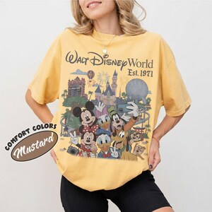 May include: A mustard yellow t-shirt featuring a graphic print of Walt Disney World Est. 1971 with Mickey Mouse, Minnie Mouse, Donald Duck, Daisy Duck, Goofy, Pluto, and Chip 'n Dale in front of the castle and Epcot ball. A Comfort Colors Mustard label.