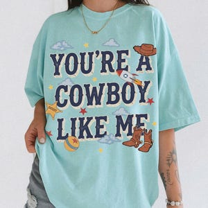 May include: A light blue t-shirt featuring the text "YOU'RE A COWBOY LIKE ME" in dark blue letters. The graphic includes a brown cowboy hat, a rocket ship, a yellow sheriff star, a colorful ball, and brown cowboy boots, surrounded by small clouds and stars.