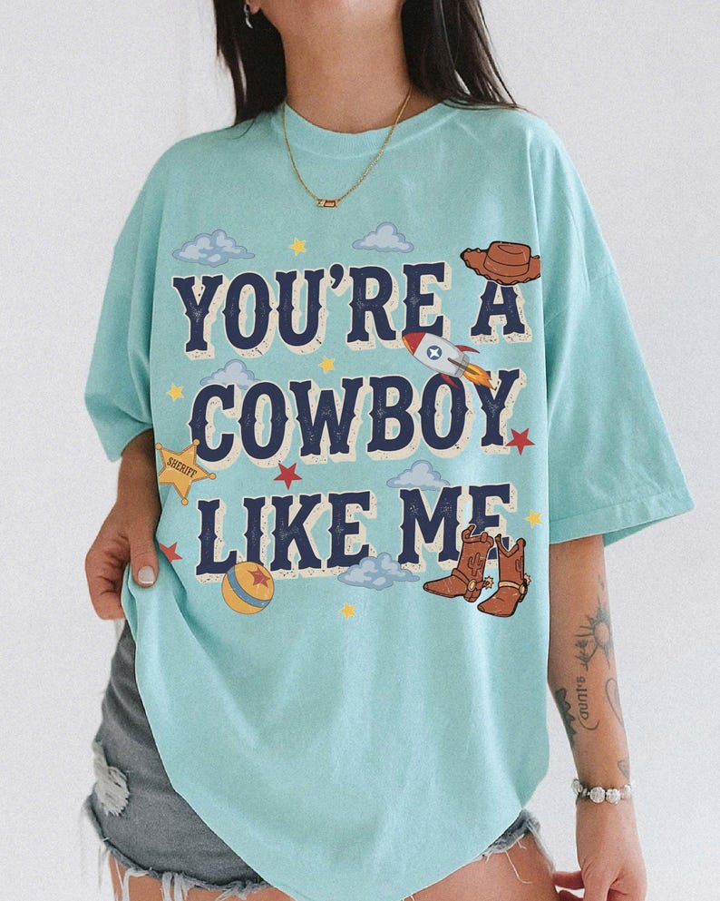 May include: A light blue t-shirt featuring the text "YOU'RE A COWBOY LIKE ME" in dark blue letters. The graphic includes a brown cowboy hat, a rocket ship, a yellow sheriff star, a colorful ball, and brown cowboy boots, surrounded by small clouds and stars.