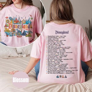 Puede incluir: Una camiseta rosa claro de Disneyland con un gr&aacute;fico colorido en la parte delantera y una lista de atracciones y fechas en la parte trasera. El gr&aacute;fico frontal presenta el logotipo de Disneyland y varios iconos del parque.