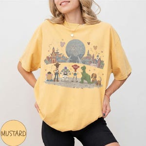 Puede incluir: Camiseta de color mostaza con un gr&aacute;fico de personajes de Toy Story frente a un parque tem&aacute;tico. El dise&ntilde;o incluye a Woody, Buzz Lightyear y otros personajes. La camiseta tiene cuello redondo y mangas cortas.