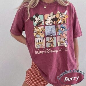 May include: Berry-colored t-shirt featuring a grid of Disney characters including Mickey Mouse, Minnie Mouse, and Goofy. The shirt has the words "Walt Disney World" printed below the grid. The shirt is labeled "Comfort Colors Berry".