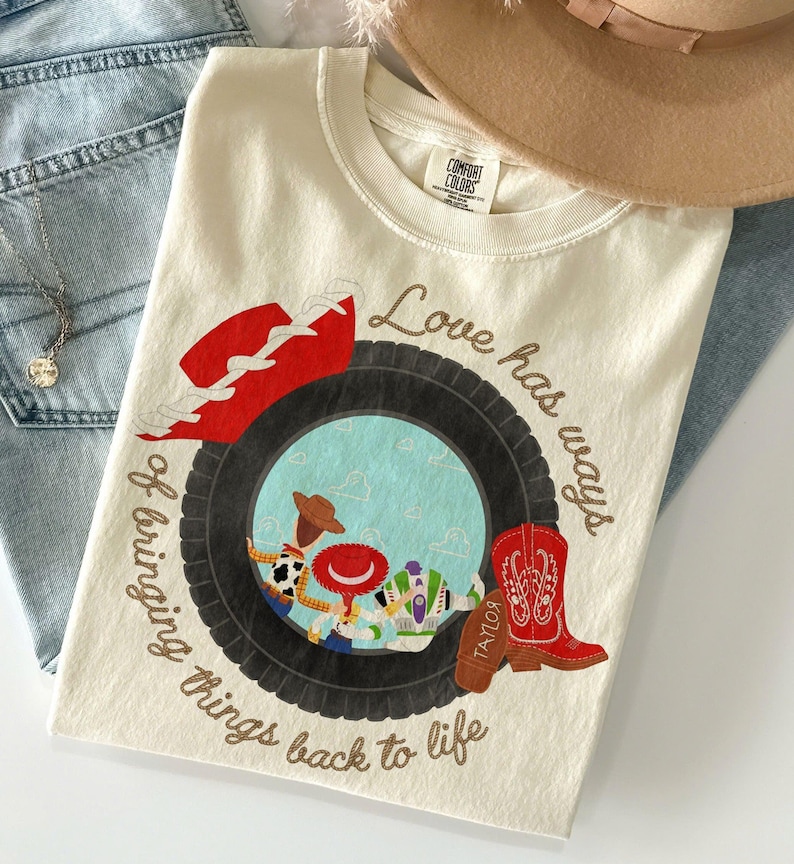 May include: A cream-colored t-shirt featuring a graphic of a black tire framing a scene with three cartoon characters. The text reads Love has ways of bringing things back to life. A red cowboy hat and a red cowboy boot with the name Taylor are included.