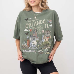 K&ouml;nnte beinhalten: Olivgr&uuml;nes T-Shirt mit einer Grafik von Wahrzeichen von Orlando, Florida, darunter ein Schloss und Feuerwerk, mit dem Text "Greetings from Orlando, FL." Das Shirt hat kurze &Auml;rmel und eine lockere Passform.