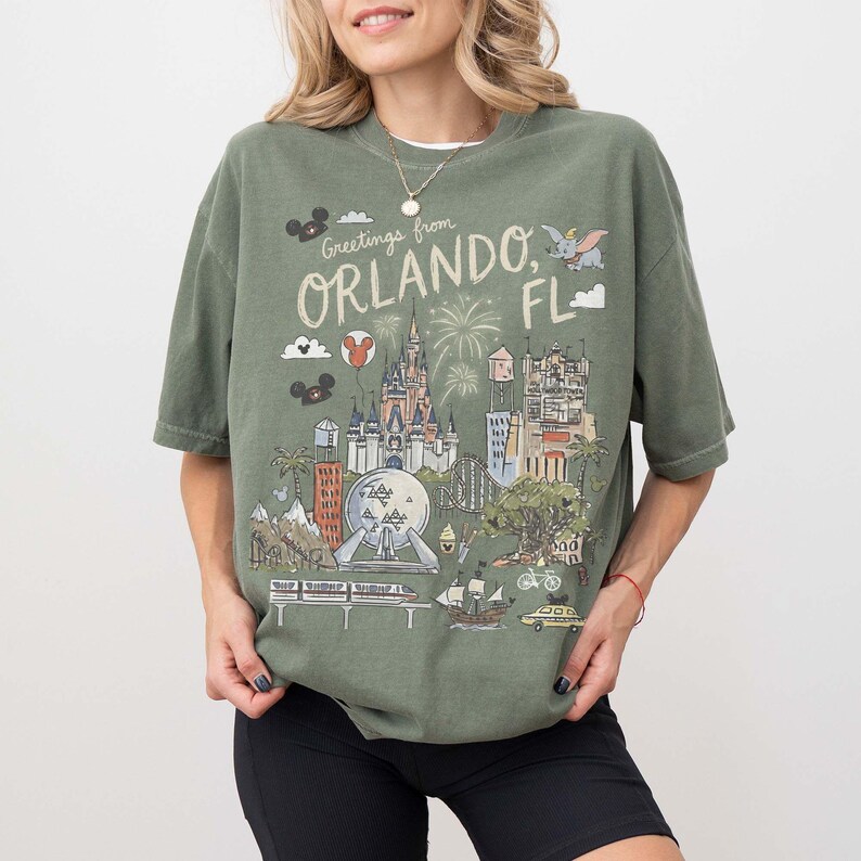 K&ouml;nnte beinhalten: Olivgr&uuml;nes T-Shirt mit einer Grafik von Wahrzeichen von Orlando, Florida, darunter ein Schloss und Feuerwerk, mit dem Text "Greetings from Orlando, FL." Das Shirt hat kurze &Auml;rmel und eine lockere Passform.
