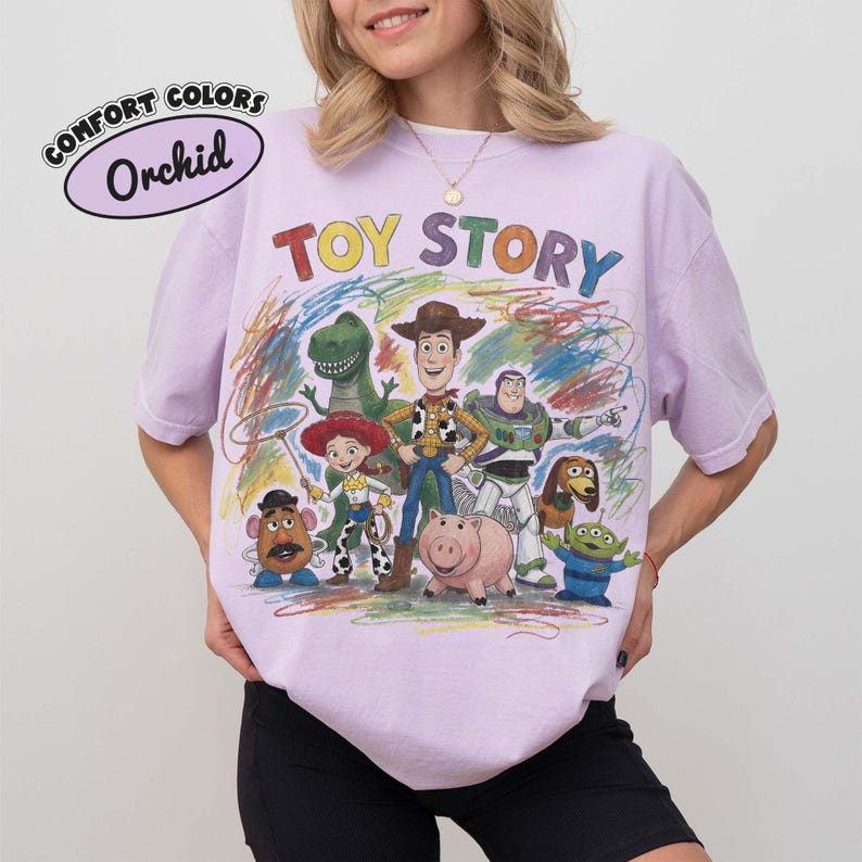 Pu&ograve; includere: Una t-shirt a maniche corte color viola chiaro con una stampa grafica colorata dei personaggi di Toy Story, tra cui Woody, Buzz Lightyear, Jessie, Rex, Slinky Dog, Mr. Potato Head, Hamm e un Alien, con il testo "TOY STORY" e "COMFORT COLORS Orchid".