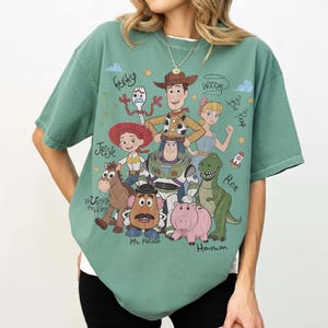 May include: A sage green short-sleeve t-shirt featuring a graphic print of Toy Story characters including Woody, Buzz Lightyear, Jessie, Rex, Hamm, Mr. Potato Head, Bullseye, Bo Peep, and Forky, with their names written in cursive script.