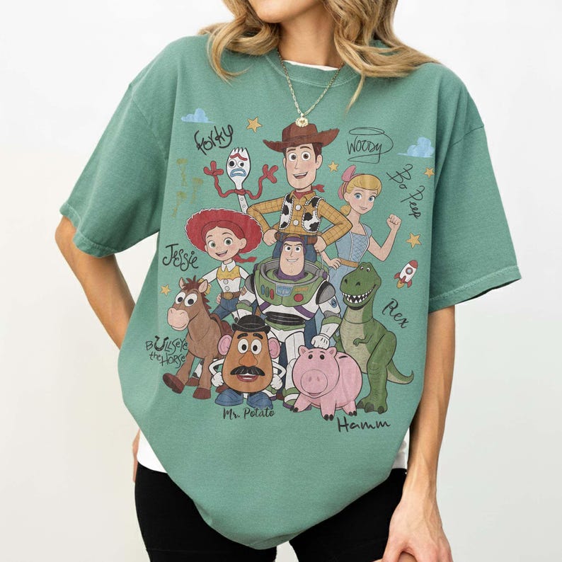 May include: A sage green short-sleeve t-shirt featuring a graphic print of Toy Story characters including Woody, Buzz Lightyear, Jessie, Rex, Hamm, Mr. Potato Head, Bullseye, Bo Peep, and Forky, with their names written in cursive script.
