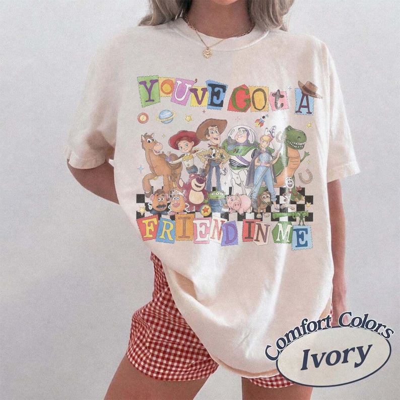May include: An ivory short-sleeve t-shirt featuring a colorful graphic print of various animated characters from the Toy Story franchise. The text "YOU'VE GOT A FRIEND IN ME" is displayed in multi-colored block letters above and below the characters.