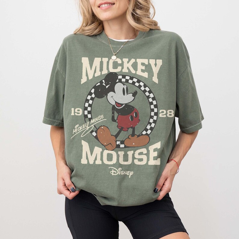 May include: Olive green t-shirt featuring a vintage Mickey Mouse graphic. The design includes Mickey Mouse in a walking pose, surrounded by a checkered circle, with the words "Mickey Mouse" and "Disney" printed on the shirt.