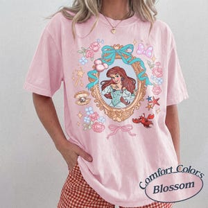 Puede incluir: Camiseta rosa claro con una ilustraci&oacute;n enmarcada de una princesa pelirroja con un vestido azul. El marco est&aacute; rodeado de elementos florales y acu&aacute;ticos, incluyendo una concha, una estrella de mar y un cangrejo rojo. Un lazo rosa en la parte inferior.