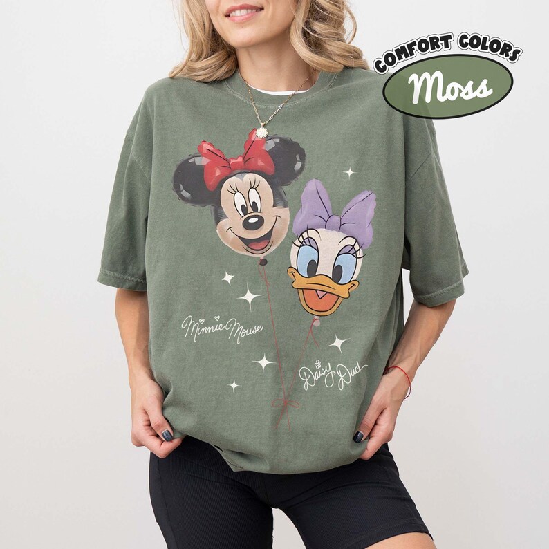 Pu&ograve; includere: Una t-shirt a maniche corte color verde muschio con una stampa grafica di Minnie Mouse e Daisy Duck come palloncini. Il design include il testo Minnie Mouse e Daisy Duck con piccole stelle. Un'etichetta nell'angolo riporta Comfort Colors Moss.