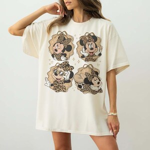 May include: Cream-colored t-shirt featuring four cartoon images of Minnie Mouse in leopard print outfits. Each image is framed by a tan-colored decorative border with small hearts and stars. The shirt has short sleeves and a relaxed fit.