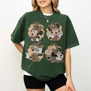 May include: Dark green t-shirt featuring four Minnie Mouse designs with leopard print accents. Each design is set within a decorative frame. The shirt has short sleeves and a relaxed fit.