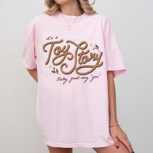 May include: A light pink short-sleeve t-shirt featuring the text "it's a Toy Story" written in a brown rope-style font, with the additional text "Baby, just say "yes"" below it, accompanied by small floral illustrations in brown and cream.