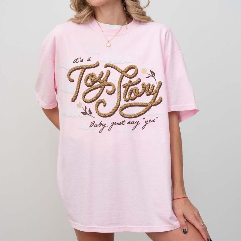 May include: A light pink short-sleeve t-shirt featuring the text "it's a Toy Story" written in a brown rope-style font, with the additional text "Baby, just say "yes"" below it, accompanied by small floral illustrations in brown and cream.