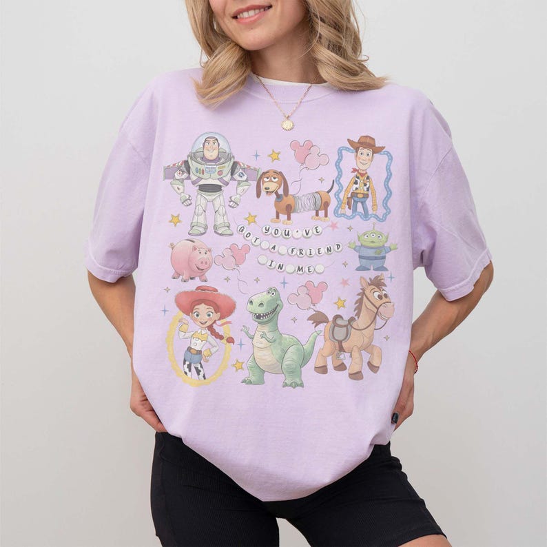 K&ouml;nnte beinhalten: Helllila T-Shirt mit Cartoon-Figuren aus dem Film Toy Story. Das Design zeigt Buzz Lightyear, Woody, Jessie und andere Charaktere. Der Text "You've Got a Friend in Me" ist auf dem Shirt aufgedruckt.