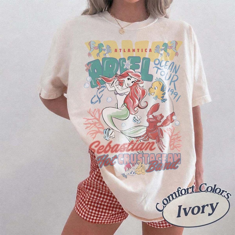 May include: Ivory-colored t-shirt featuring a graphic of Ariel from The Little Mermaid, with the text "Ocean Tour 1991." The design includes illustrations of Ariel, Flounder, and Sebastian. The shirt is labeled "Comfort Colors."