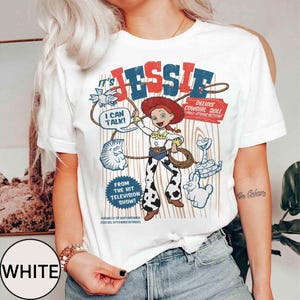 May include: White t-shirt with a cartoon graphic of Jessie from Toy Story. The design includes the text "JESSIE" in red and blue, along with other cartoon characters and text bubbles. The shirt also has the text "DELUXE COWGIRL DOLL" and "FROM THE HIT TELEVISION SHOW!"
