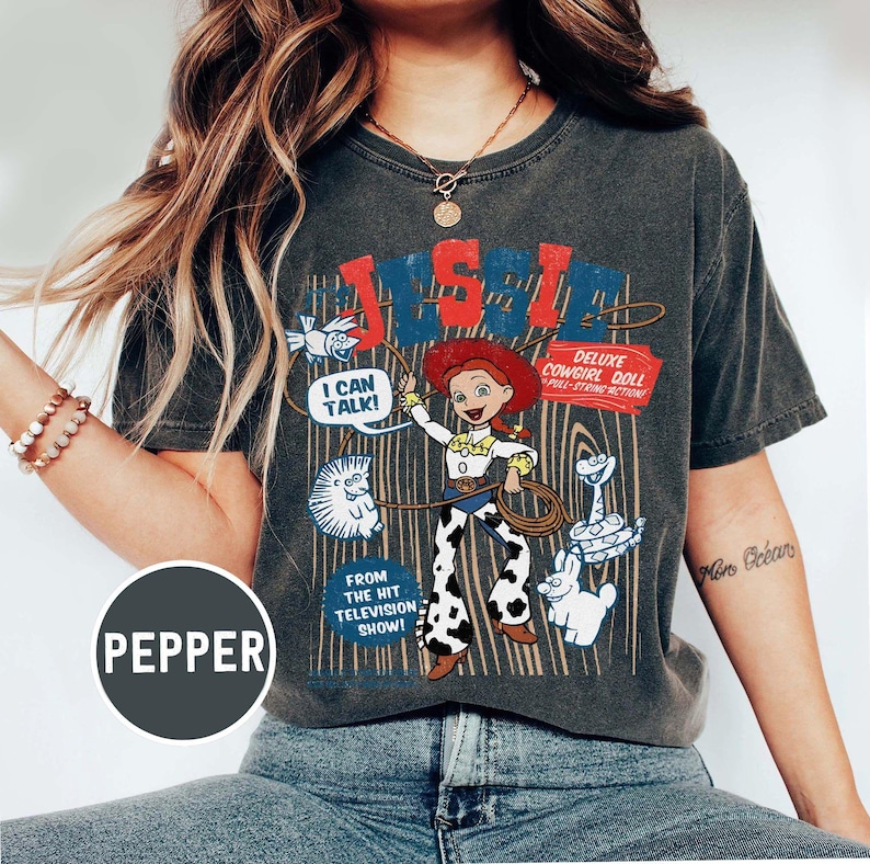 May include: Dark grey t-shirt with a graphic of a cowgirl doll and the text "JESSIE" in red and blue. The design includes the text "I CAN TALK!" and "DELUXE COWGIRL DOLL". The shirt is from the hit television show.