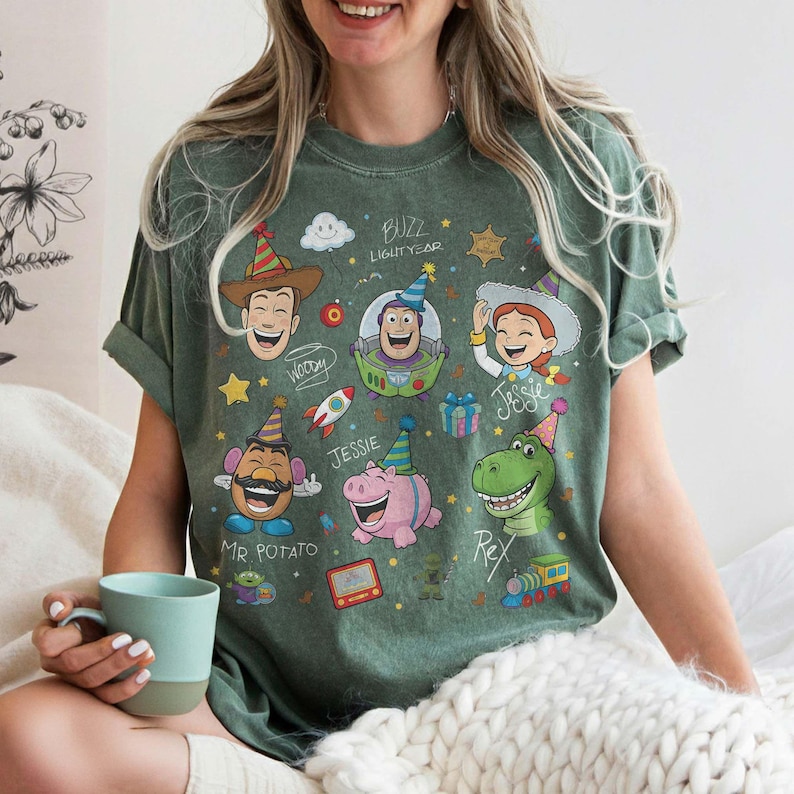 May include: Green t-shirt featuring cartoon illustrations of characters from the movie Toy Story, including Woody, Buzz Lightyear, Jessie, Mr. Potato Head, and Rex. The shirt has a crew neck and short sleeves.