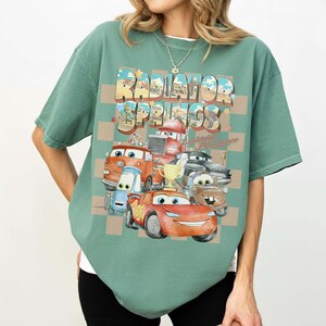 Peut inclure: T-shirt vert avec un motif r&eacute;tro de voitures anim&eacute;es et le texte "Radiator Springs". Le design pr&eacute;sente divers personnages de voitures et un fond &agrave; carreaux, offrant une esth&eacute;tique ludique et nostalgique.