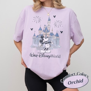 May include: A lilac t-shirt with a graphic of Mickey Mouse in front of a castle and fireworks. The text "Walt Disney World" is printed below the graphic. The shirt is labelled "Comfort Colors Orchid".