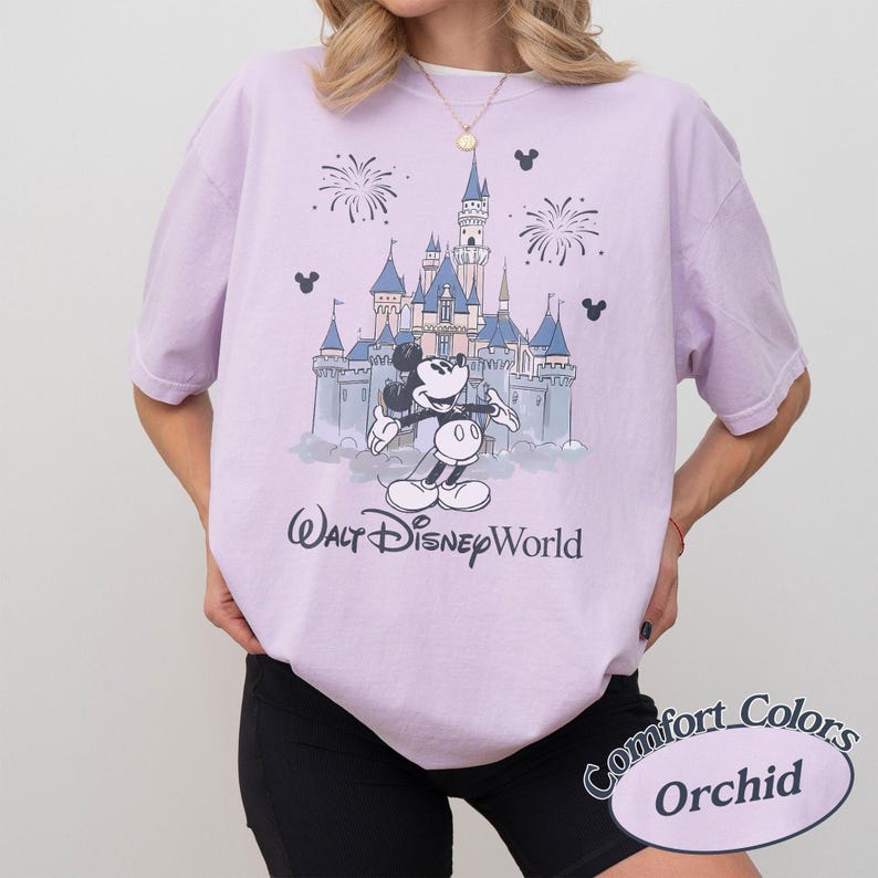 May include: A lilac t-shirt with a graphic of Mickey Mouse in front of a castle and fireworks. The text "Walt Disney World" is printed below the graphic. The shirt is labelled "Comfort Colors Orchid".