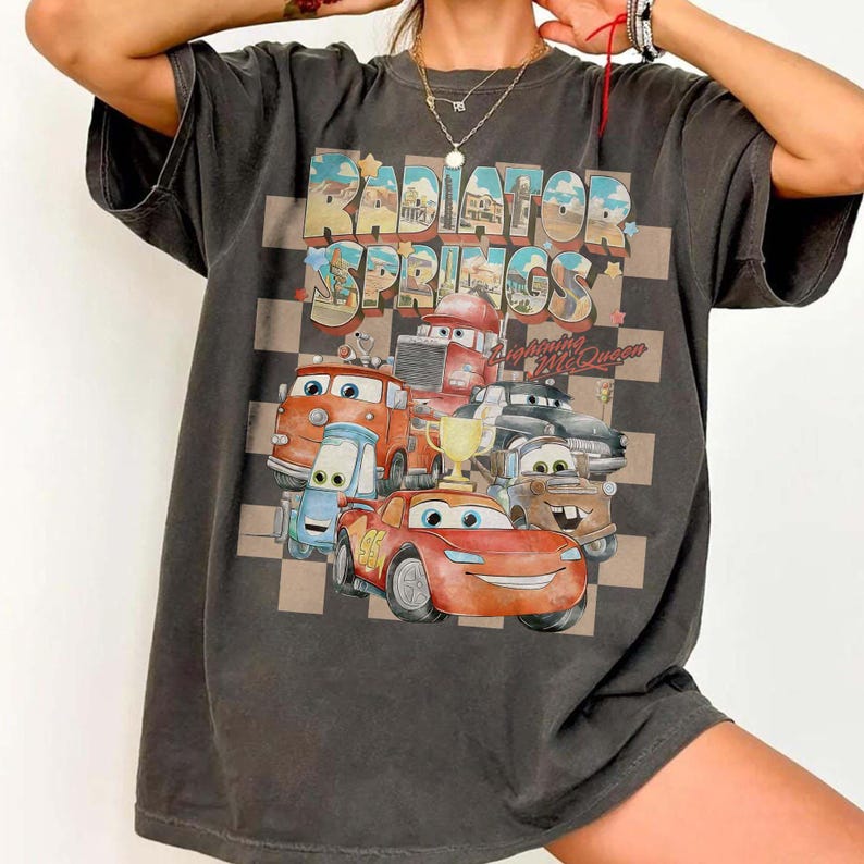 Peut inclure: T-shirt gris fonc&eacute; avec un graphisme color&eacute; des personnages du film Cars, dont Flash McQueen, et le texte "Radiator Springs" dans un style vintage. Le motif est plac&eacute; sur un fond &agrave; carreaux.