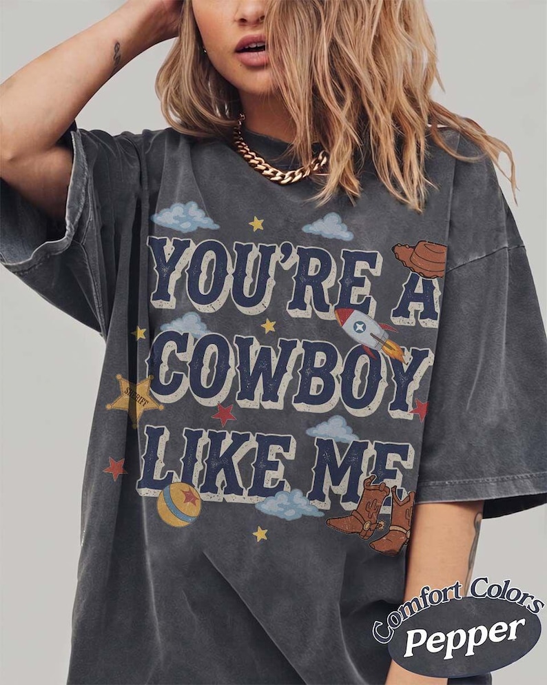 May include: A charcoal gray oversized t-shirt featuring the text "YOU'RE A COWBOY LIKE ME" in dark blue block letters. The graphic includes illustrations of a rocket, a cowboy hat, a sheriff star, a ball, and cowboy boots with small clouds and stars.