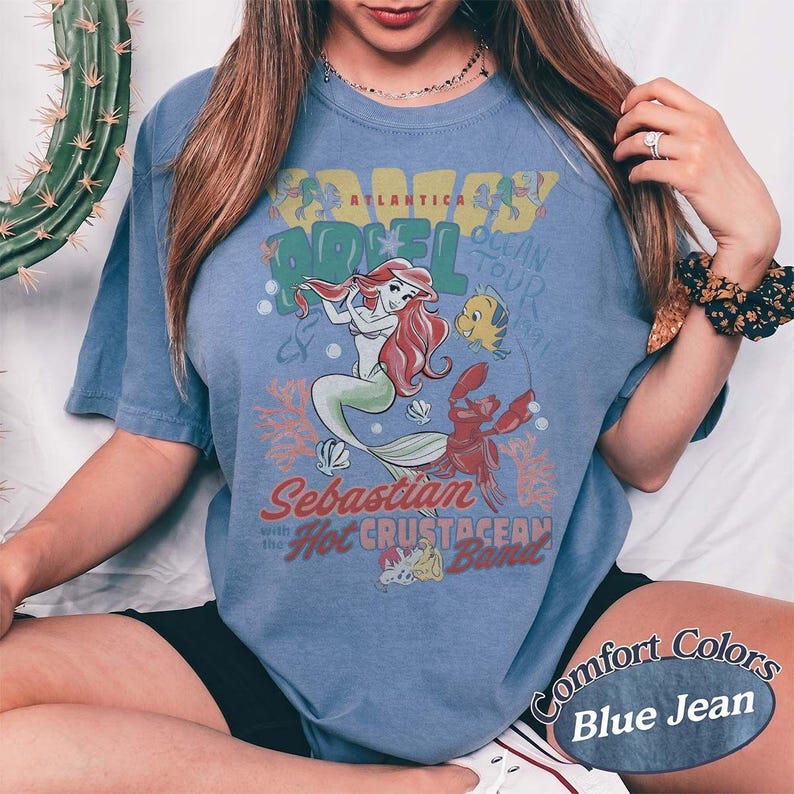 May include: Blue jean colored t-shirt featuring a graphic of Ariel from The Little Mermaid, with Sebastian and Flounder. The design includes the text "Crazy Ariel Ocean Tour" and "Sebastian with the Hot Crustacean Band."