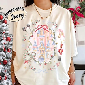 May include: Ivory Comfort Colors t-shirt featuring a whimsical Christmas castle design. The design includes a castle, a wreath, a toy soldier, and Mickey Mouse ears. The shirt has the word "Ivory" in a circular design.