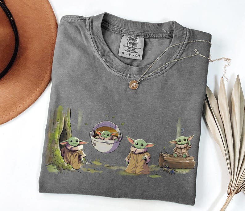 May include: A charcoal gray short-sleeve t-shirt featuring a graphic print of four small green creatures with large ears in various poses, including one in a floating pod. The shirt has a Comfort Colors brand label at the neck indicating size S.