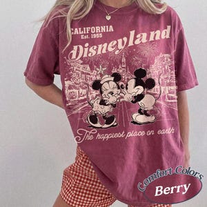 Puede incluir: Camiseta de color bayas con un gr&aacute;fico de Disneyland con Mickey y Minnie Mouse. La camiseta tiene el texto "CALIFORNIA Est. 1955" y "The happiest place on earth". La camiseta es de la marca Comfort Colors.