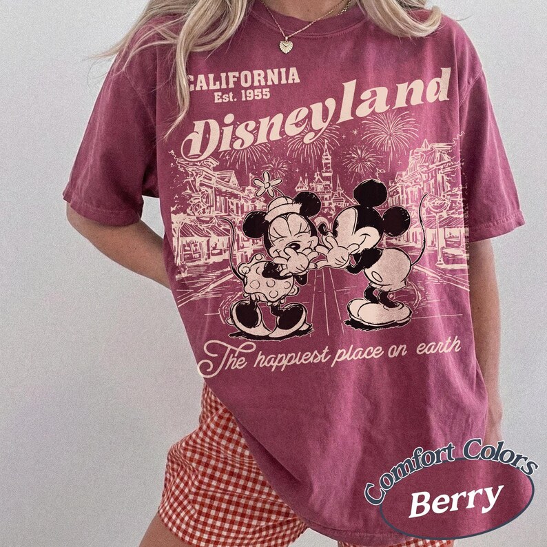 Puede incluir: Camiseta de color bayas con un gr&aacute;fico de Disneyland con Mickey y Minnie Mouse. La camiseta tiene el texto "CALIFORNIA Est. 1955" y "The happiest place on earth". La camiseta es de la marca Comfort Colors.