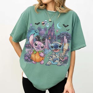 May include: A sage green short-sleeve t-shirt featuring a graphic print of two blue alien characters in Halloween costumes. One character wears a pumpkin suit, the other is wrapped in bandages like a mummy, set against a theme park castle background.
