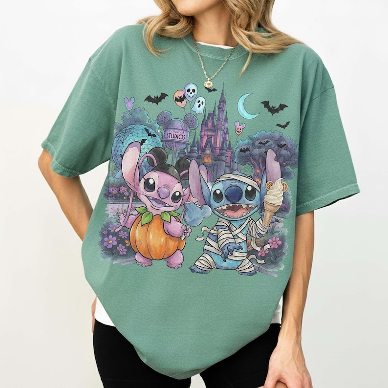 May include: A sage green short-sleeve t-shirt featuring a graphic print of two blue alien characters in Halloween costumes. One character wears a pumpkin suit, the other is wrapped in bandages like a mummy, set against a theme park castle background.