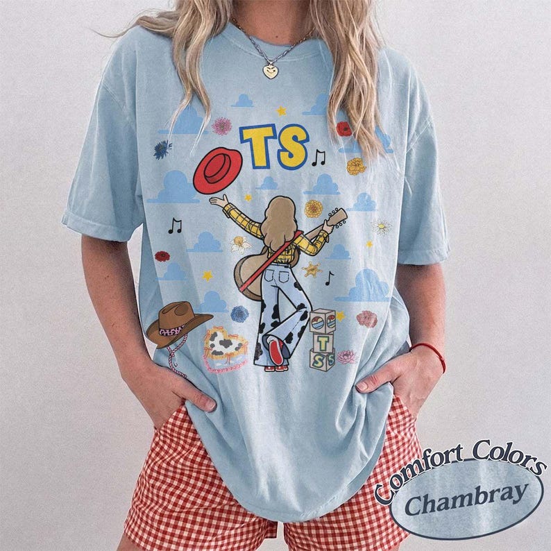 May include: A light blue t-shirt featuring a graphic print of a person with long hair playing a guitar, wearing cow-print pants. The design includes the text TS, a red hat, a brown hat, musical notes, flowers, clouds, and stacked blocks with TS.