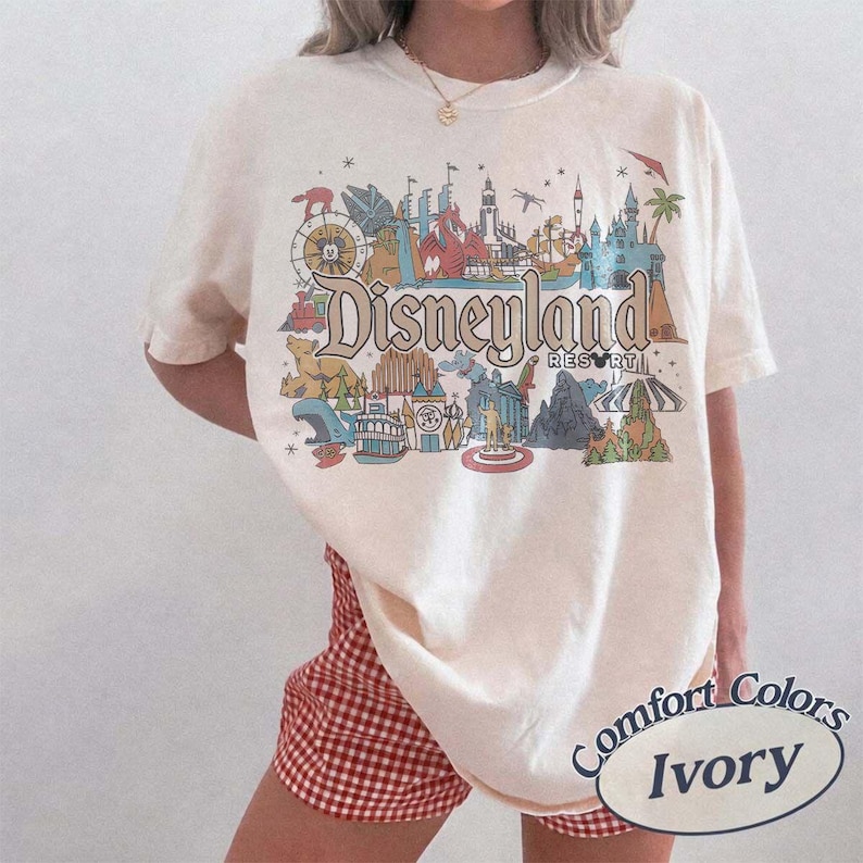 May include: Ivory-colored t-shirt featuring a colorful Disneyland Resort graphic. The design includes iconic park landmarks and the Disneyland logo. The shirt is a Comfort Colors brand, with the color name "Ivory" printed on the bottom right.
