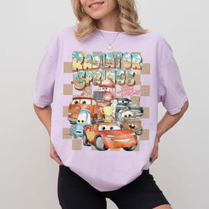 Peut inclure: T-shirt lilas clair avec un motif des personnages du film Cars. Le motif comprend le texte "Radiator Springs" et "Lightning McQueen" avec des images des personnages. Le t-shirt a un col rond et des manches courtes.