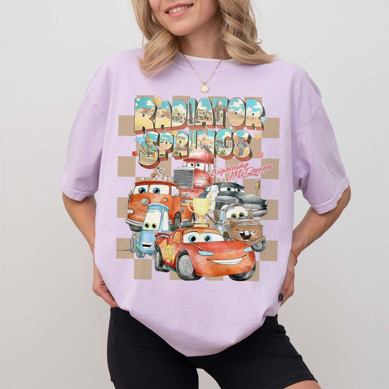 Peut inclure: T-shirt lilas clair avec un motif des personnages du film Cars. Le motif comprend le texte "Radiator Springs" et "Lightning McQueen" avec des images des personnages. Le t-shirt a un col rond et des manches courtes.