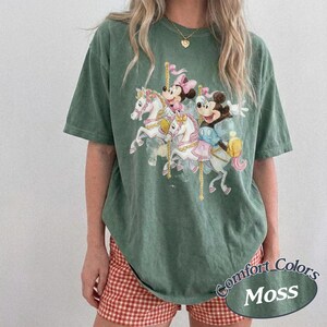 Op de afbeelding: Groen T-shirt met een carrouselontwerp met Mickey en Minnie Mouse. Het shirt heeft een losse pasvorm en de woorden "Comfort Colors Moss" staan rechtsonder gedrukt. Het model draagt een rood-wit geruite short.