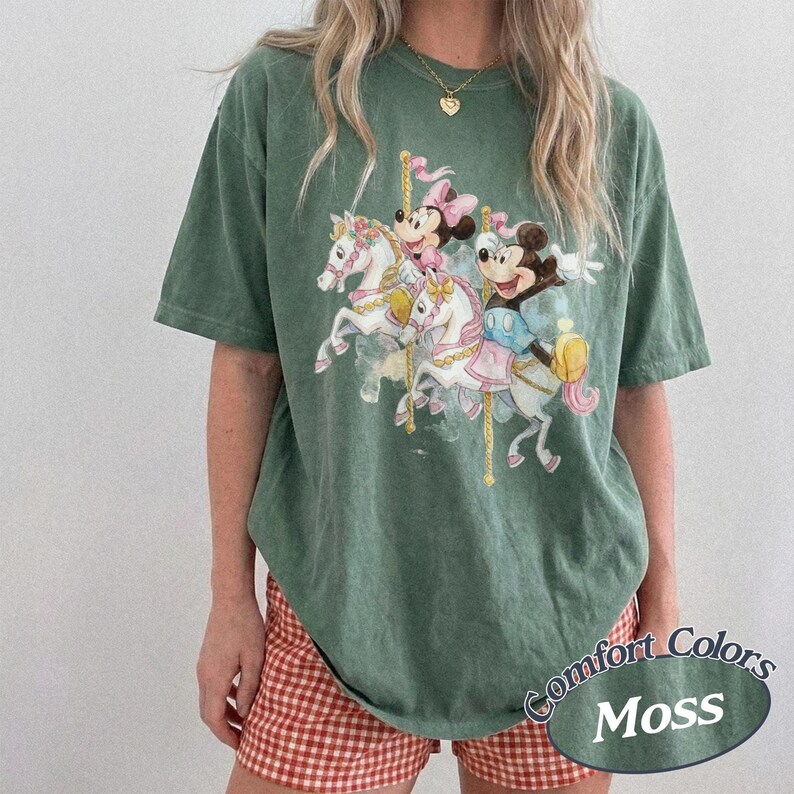 Op de afbeelding: Groen T-shirt met een carrouselontwerp met Mickey en Minnie Mouse. Het shirt heeft een losse pasvorm en de woorden "Comfort Colors Moss" staan rechtsonder gedrukt. Het model draagt een rood-wit geruite short.