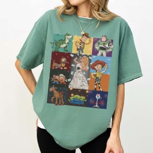May include: A sage green short-sleeve t-shirt featuring a grid of Toy Story characters including Rex, Woody, Buzz Lightyear, Slinky Dog, Mr. Potato Head, Jessie, Bullseye, the Aliens, and Forky, surrounding a central illustration of a singer.