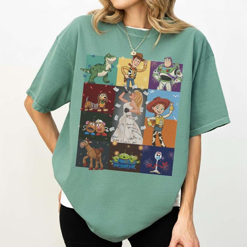 May include: A sage green short-sleeve t-shirt featuring a grid of Toy Story characters including Rex, Woody, Buzz Lightyear, Slinky Dog, Mr. Potato Head, Jessie, Bullseye, the Aliens, and Forky, surrounding a central illustration of a singer.